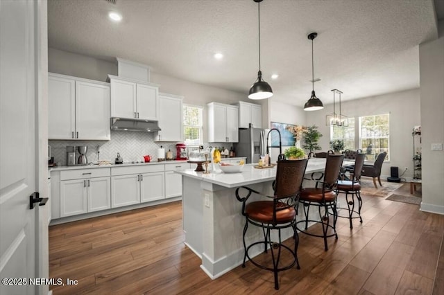 kitchen with wood finished floors, an island with sink, stainless steel fridge with ice dispenser, light countertops, and under cabinet range hood