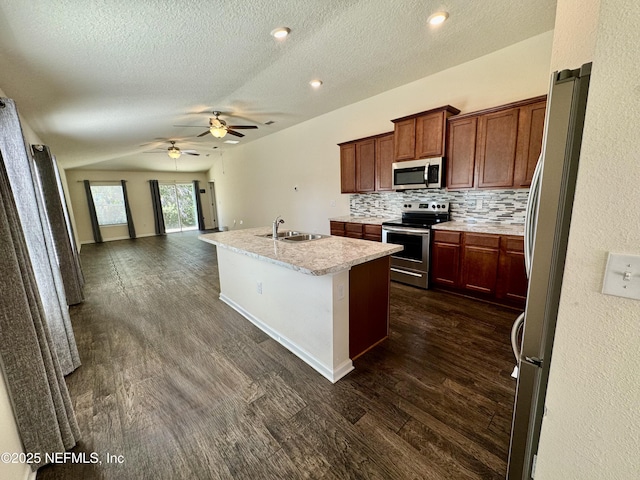 kitchen with a kitchen island with sink, dark wood-style flooring, a sink, decorative backsplash, and appliances with stainless steel finishes