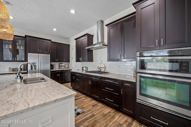 kitchen featuring light stone counters, a sink, wood finished floors, appliances with stainless steel finishes, and wall chimney range hood