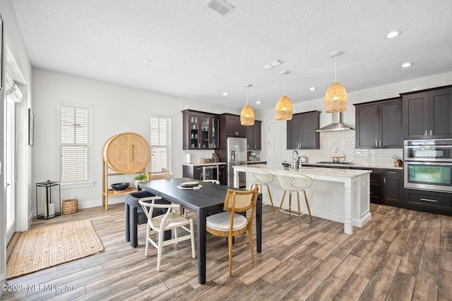 kitchen with wall chimney exhaust hood, dark wood-style floors, stainless steel appliances, a kitchen island with sink, and visible vents