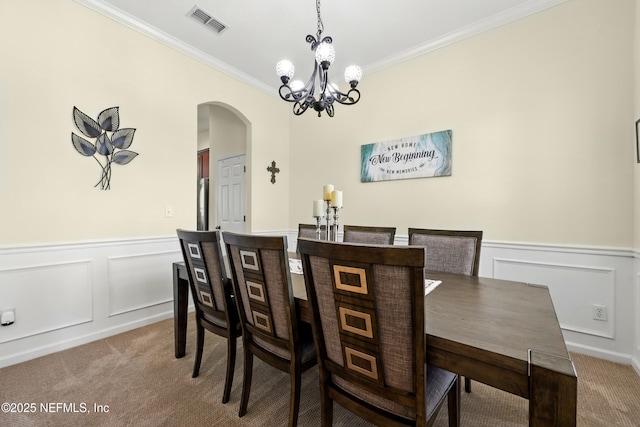 carpeted dining room with arched walkways, a wainscoted wall, crown molding, a decorative wall, and a chandelier