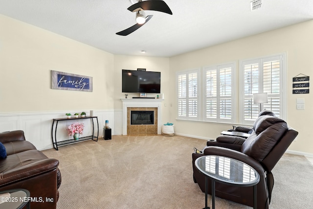 living area with ceiling fan, carpet, a tile fireplace, and a wainscoted wall