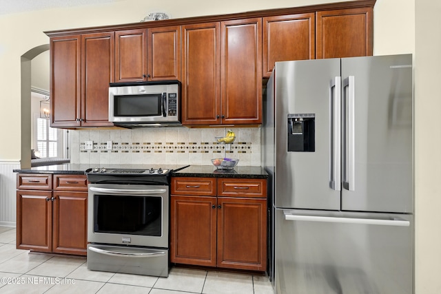 kitchen with appliances with stainless steel finishes, dark stone counters, light tile patterned floors, and decorative backsplash