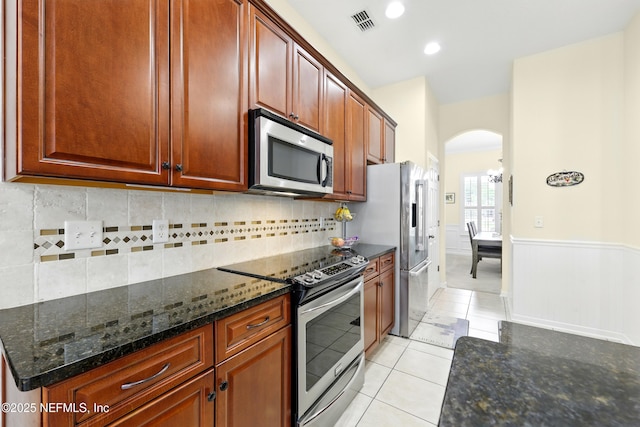 kitchen with stainless steel appliances, arched walkways, backsplash, light tile patterned floors, and dark stone countertops
