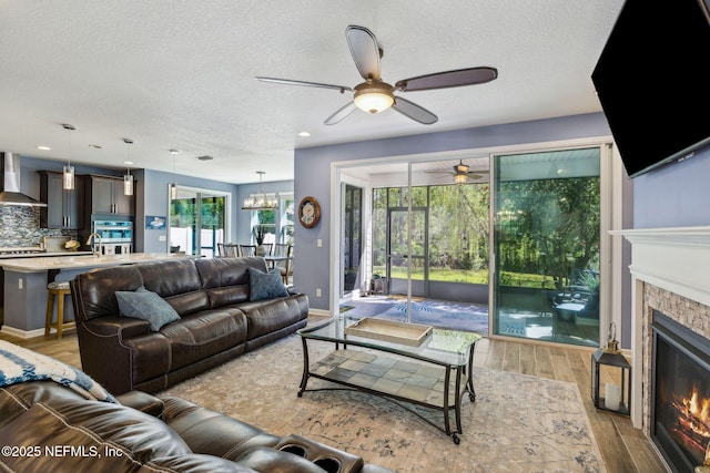 living room featuring a textured ceiling, baseboards, light wood-style floors, ceiling fan with notable chandelier, and a glass covered fireplace
