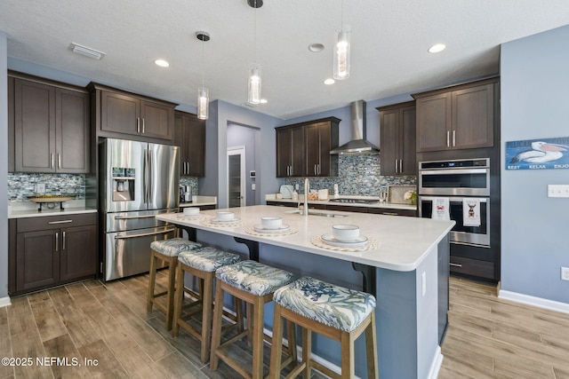 kitchen featuring light countertops, wall chimney exhaust hood, a kitchen bar, appliances with stainless steel finishes, and dark brown cabinets
