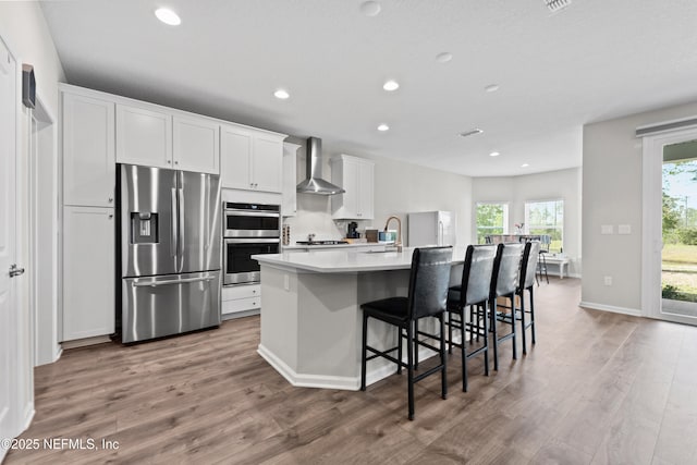 kitchen with stainless steel appliances, wall chimney range hood, white cabinets, a kitchen island with sink, and wood finished floors
