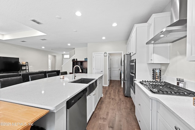 kitchen with a sink, white cabinetry, appliances with stainless steel finishes, wall chimney range hood, and a kitchen island with sink