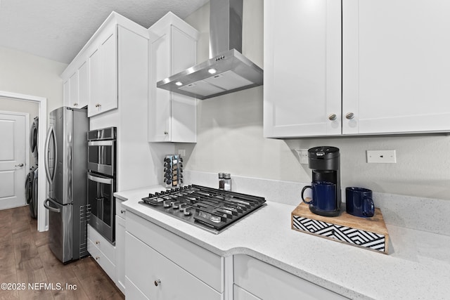 kitchen featuring wall chimney range hood, white cabinetry, stacked washing maching and dryer, and stainless steel appliances
