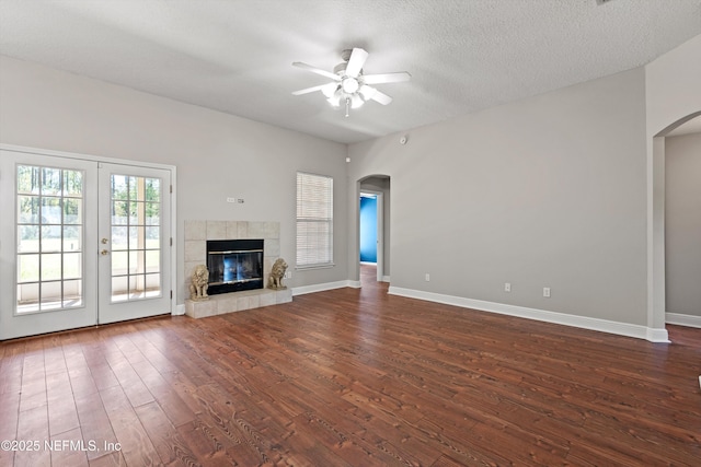 unfurnished living room featuring baseboards, arched walkways, dark wood-style floors, ceiling fan, and a tiled fireplace