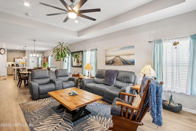 living room featuring light wood-style flooring, plenty of natural light, ceiling fan with notable chandelier, and a raised ceiling