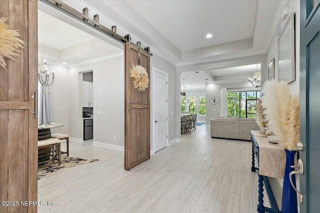 corridor featuring light wood-style floors, baseboards, a barn door, a chandelier, and a tray ceiling