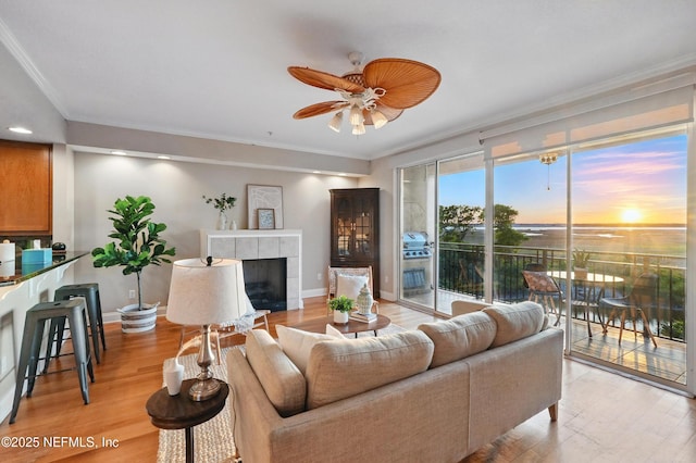 living area with light wood-type flooring, a tile fireplace, a ceiling fan, and ornamental molding