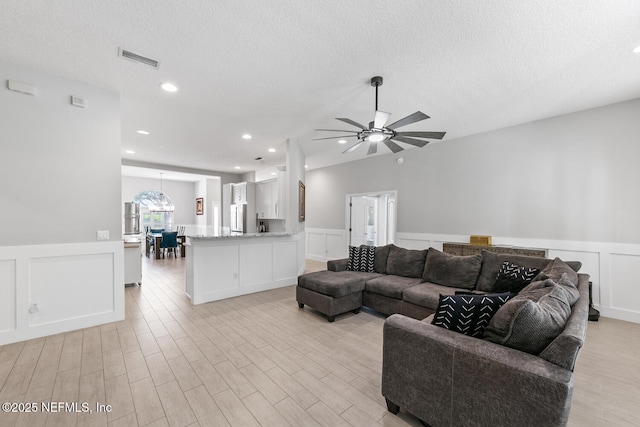 living area with light wood-style floors, a decorative wall, a textured ceiling, a ceiling fan, and visible vents