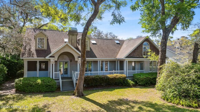 new england style home with a chimney, covered porch, a shingled roof, a front yard, and stucco siding