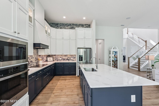 kitchen featuring black electric cooktop, a sink, a center island with sink, stainless steel oven, and blue cabinetry