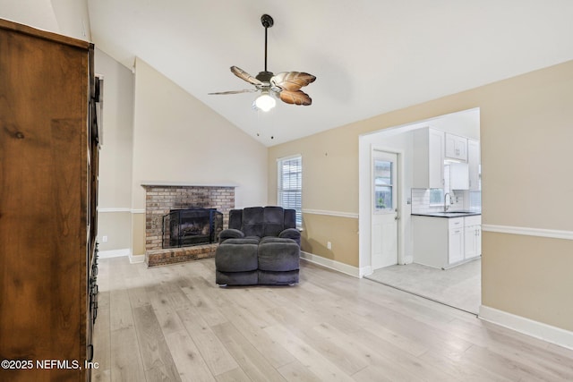 sitting room featuring a ceiling fan, a brick fireplace, light wood finished floors, vaulted ceiling, and baseboards