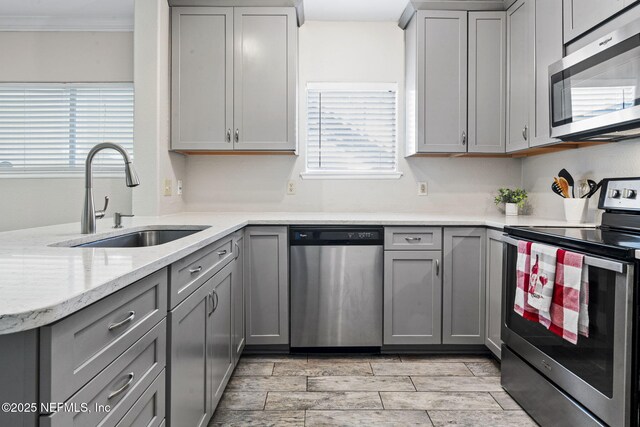 kitchen with gray cabinetry, light stone counters, a sink, ornamental molding, and stainless steel appliances