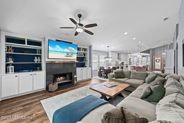 living room featuring built in features, ceiling fan, dark wood-type flooring, a fireplace with flush hearth, and visible vents