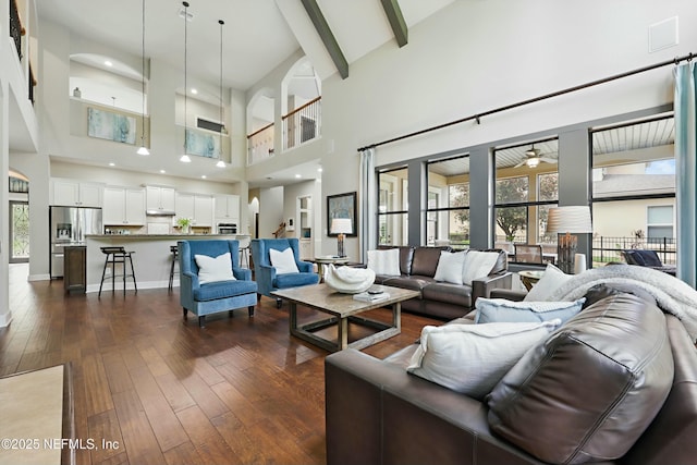 living area with a towering ceiling, dark wood-style flooring, a ceiling fan, and beam ceiling