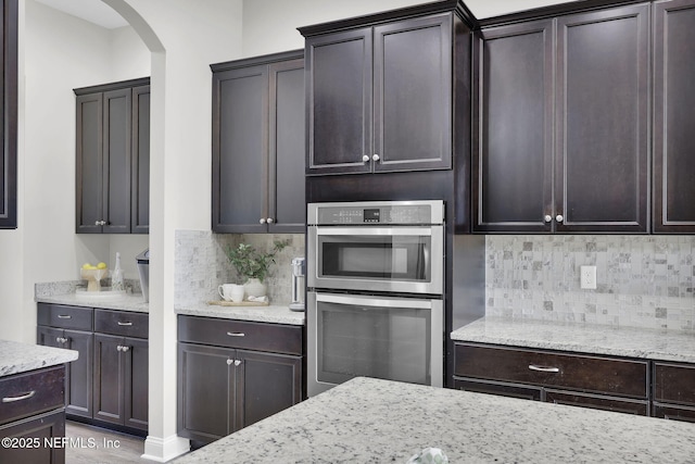 kitchen featuring tasteful backsplash, light stone counters, dark brown cabinets, stainless steel double oven, and arched walkways