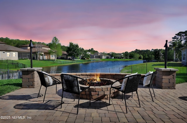 patio terrace at dusk featuring a water view, a yard, a fire pit, and fence
