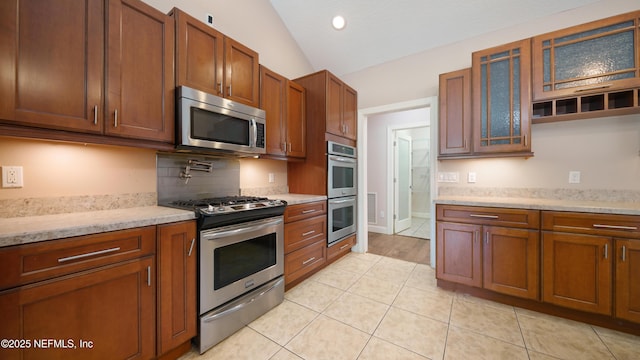 kitchen featuring light tile patterned floors, glass insert cabinets, brown cabinets, lofted ceiling, and appliances with stainless steel finishes