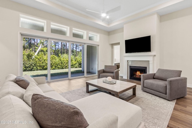 living area featuring a tray ceiling, a ceiling fan, a warm lit fireplace, wood finished floors, and baseboards