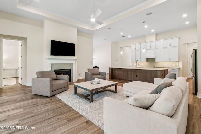 living area with baseboards, a raised ceiling, light wood-type flooring, a ceiling fan, and a lit fireplace