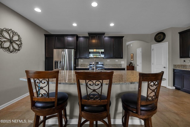 kitchen featuring light stone counters, arched walkways, appliances with stainless steel finishes, and an island with sink