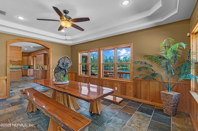 dining area with a wainscoted wall, a tray ceiling, stone tile flooring, and ceiling fan