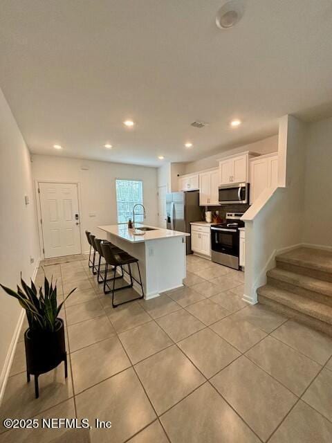 kitchen with stainless steel appliances, a sink, light tile patterned flooring, a center island with sink, and white cabinetry