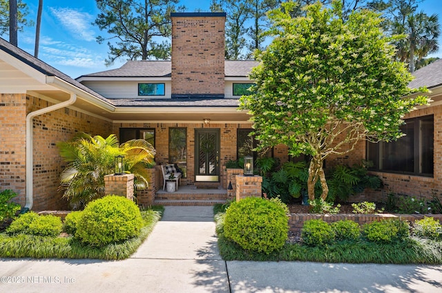 entrance to property with covered porch, brick siding, a chimney, and roof with shingles