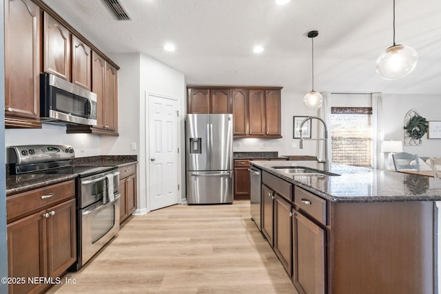 kitchen with light wood-style flooring, dark stone countertops, appliances with stainless steel finishes, a sink, and hanging light fixtures