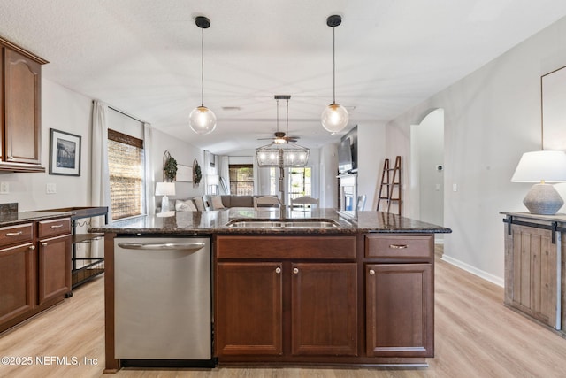 kitchen with stainless steel dishwasher, an island with sink, light wood-style flooring, a ceiling fan, and dark stone countertops