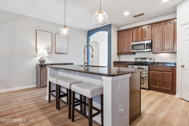kitchen featuring arched walkways, a sink, light wood-style floors, a center island with sink, and stainless steel appliances