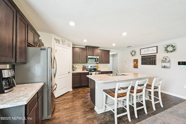 kitchen with a center island with sink, dark wood finished floors, dark brown cabinets, a sink, and stainless steel appliances