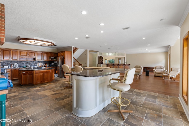 kitchen with a breakfast bar, visible vents, a center island with sink, brown cabinetry, and stainless steel refrigerator with ice dispenser
