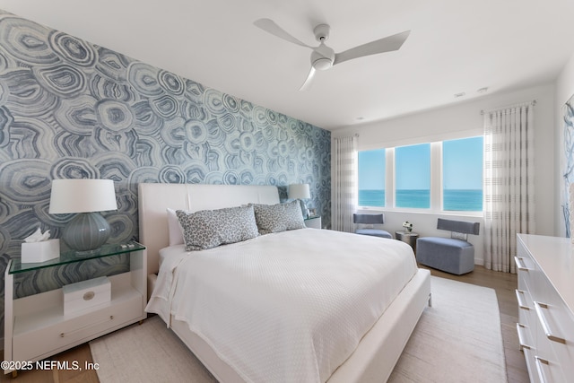 bedroom with wood finished floors, an accent wall, ceiling fan, wallpapered walls, and a water view