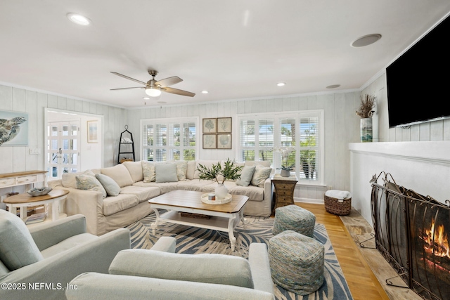 living area featuring a warm lit fireplace, ceiling fan, light wood-type flooring, wooden walls, and ornamental molding