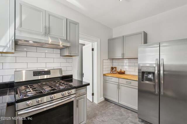 kitchen featuring butcher block counters, stainless steel appliances, backsplash, under cabinet range hood, and gray cabinetry