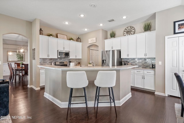 kitchen with a kitchen island with sink, arched walkways, dark wood-style flooring, backsplash, and appliances with stainless steel finishes