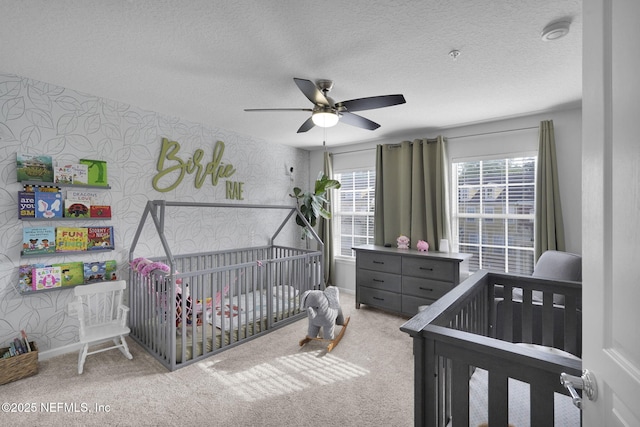 bedroom featuring a textured ceiling, wallpapered walls, ceiling fan, carpet floors, and a nursery area