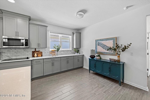 kitchen featuring stainless steel microwave, baseboards, backsplash, gray cabinetry, and black electric stovetop