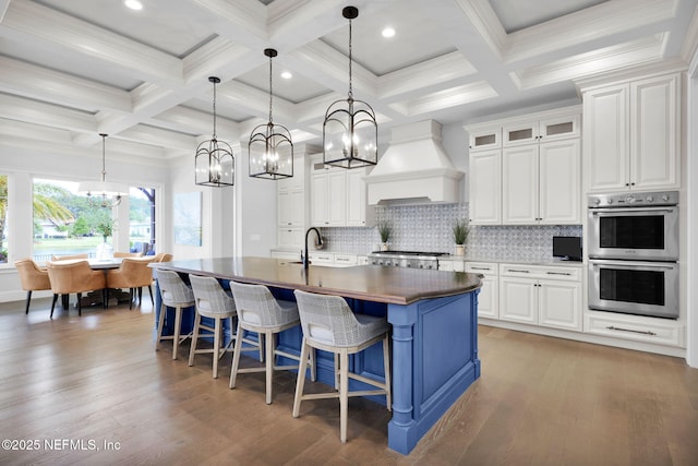 kitchen featuring an island with sink, premium range hood, stainless steel double oven, a chandelier, and light wood-style flooring