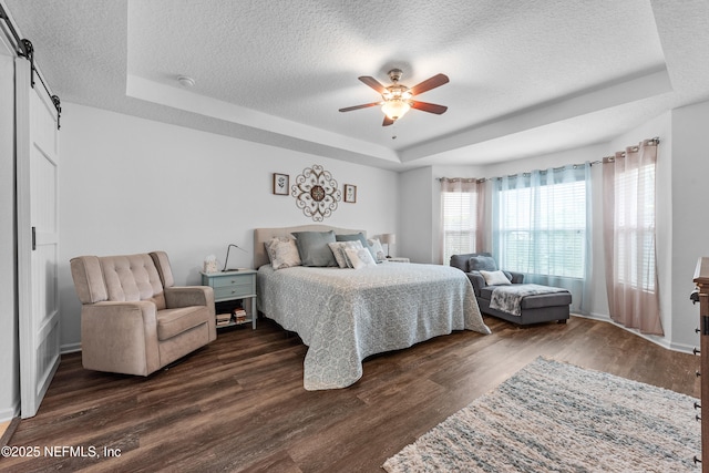 bedroom with ceiling fan, a textured ceiling, a barn door, dark wood-style floors, and a tray ceiling