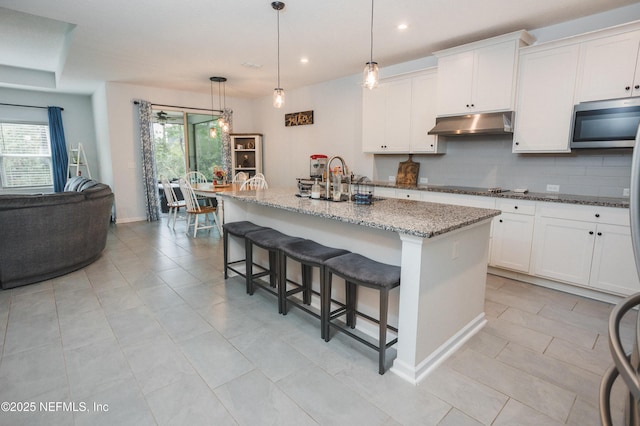 1209 Hammock Dunes Dr, Jacksonville, FL 32221 - kitchen with stainless steel microwave, white cabinetry, under cabinet range hood, light stone countertops, and a kitchen island with sink