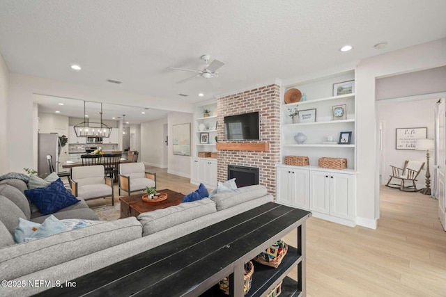 living room with light wood-style floors, a brick fireplace, a textured ceiling, and ceiling fan with notable chandelier