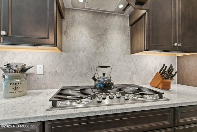 kitchen featuring dark brown cabinets, light stone countertops, stainless steel gas cooktop, decorative backsplash, and range hood