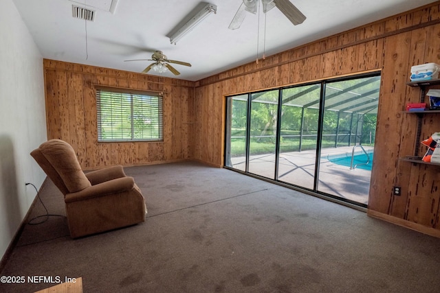 unfurnished living room featuring a ceiling fan, visible vents, wood walls, baseboards, and carpet floors